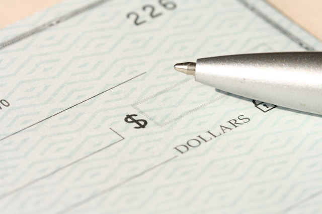 A close-up image of a blank check with a security pattern, showing the dollar sign box and part of the “DOLLARS” line. A metallic ballpoint pen rests near the writing area